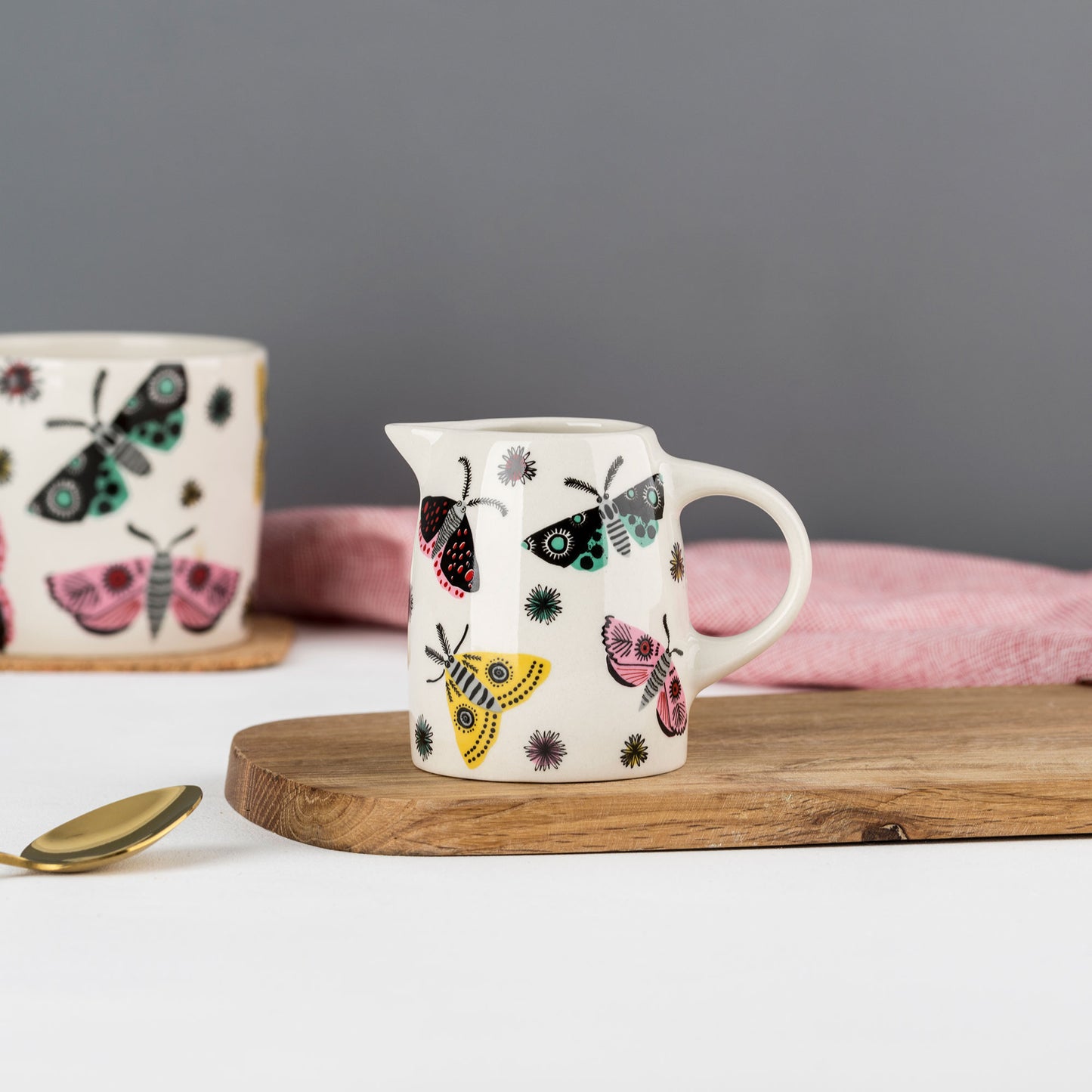 A ceramic jug with a moth pattern, placed on a wooden board next to a pink cloth, with a cup in the background that also features a moth design.