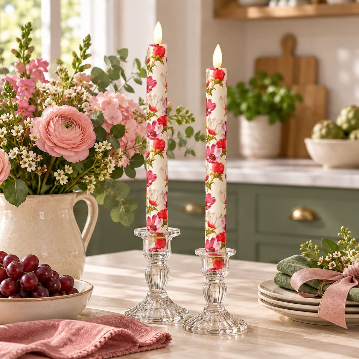 Two tall LED, fabric wrapped taper candles with a red and pink floral pattern, displayed in clear glass candle holders on a kitchen table. The scene is styled with a large vase of pink flowers, a bowl of red grapes, and folded pink napkins.