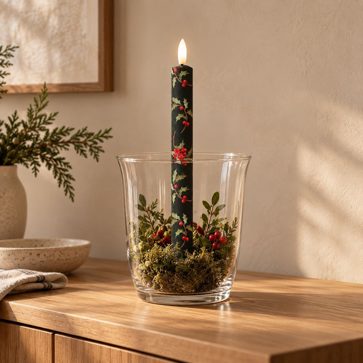 A lit LED taper candle wrapped in a dark green holly and berry print, standing upright in a clear glass vessel filled with moss and small greenery, placed on a wooden table in a warm indoor setting.