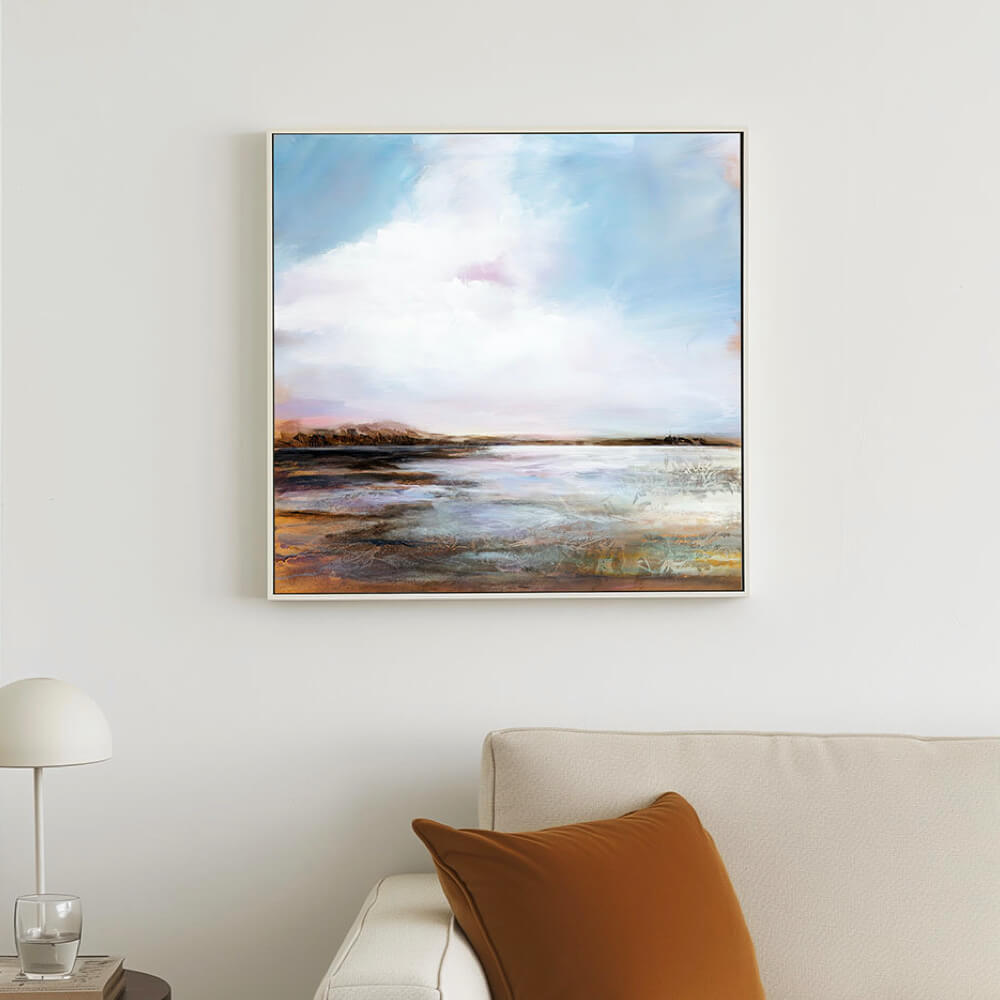 A framed canvas print of a serene ocean landscape with a dramatic sky, displayed on a wall above a white sofa with a brown cushion.