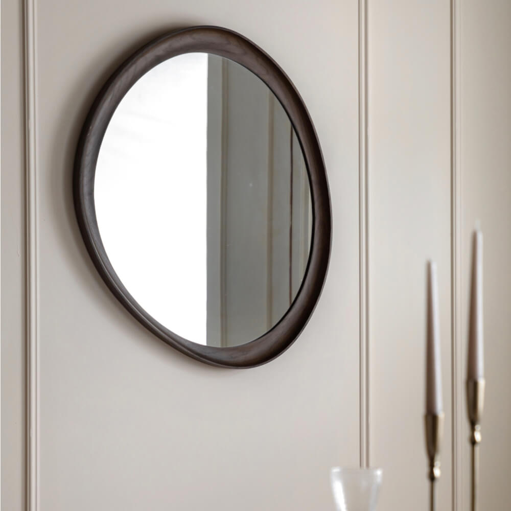 An oval wall mirror with a walnut brown wooden frame mounted on a beige panelled wall, with decorative candlesticks partially visible on the right.