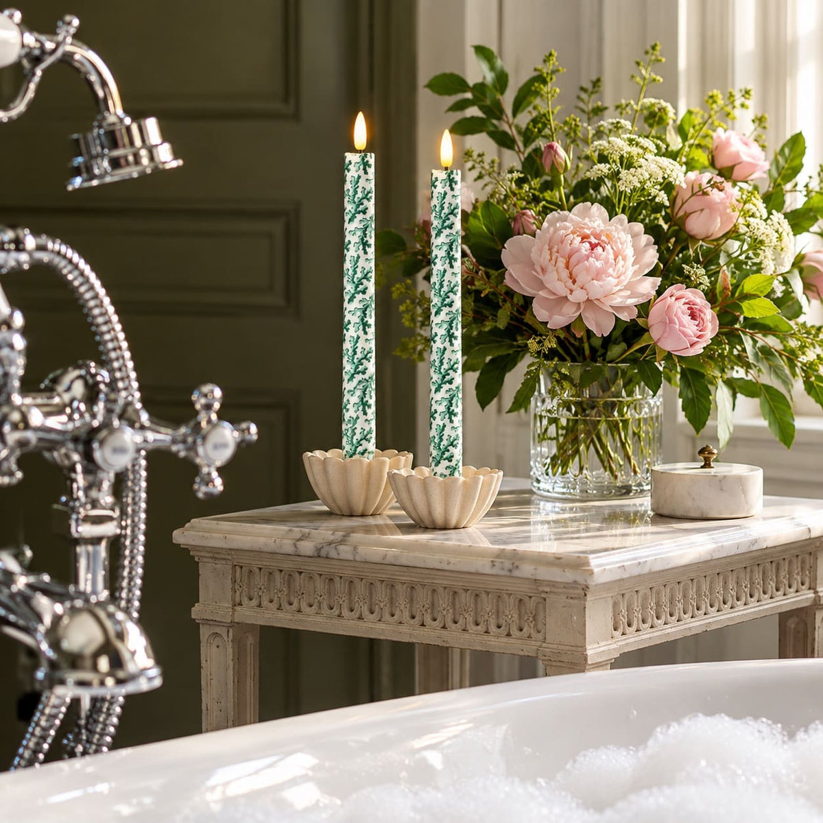 A styled bathroom scene with a marble side table holding two green coral patterned fabric LED candles, a glass vase of pink peonies, and a small white box, next to a chrome tub faucet and a bubble-filled bathtub.