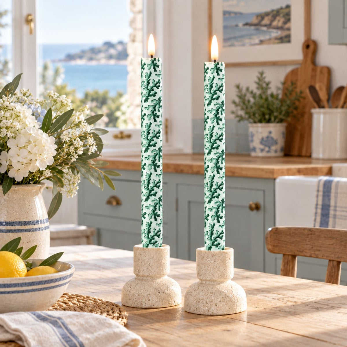 Two lit LED taper candles with a green and white coral pattern, set in round textured holders on a wooden dining table in a bright kitchen with a window view of the sea.