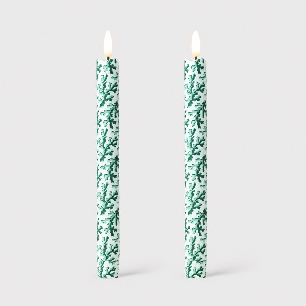 Two tall taper LED candles with a green coral pattern on fabric, shown lit against a light grey studio background.