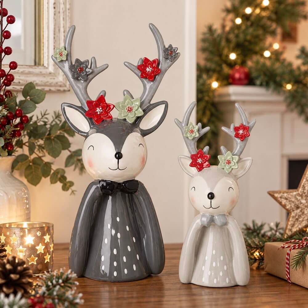 Two ceramic Christmas deer figurines with antlers decorated with red and green floral accents, displayed on a wooden table in a cozy holiday setting with lights and pine decor.