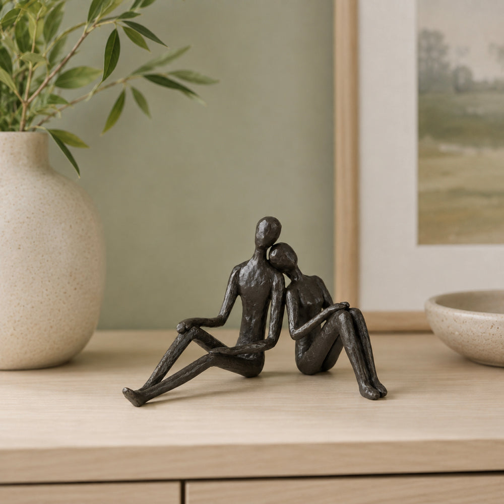 A dark bronze-finished decorative sculpture of a seated couple, displayed on a light wood console table in a styled interior setting with a ceramic vase and framed artwork in the background.