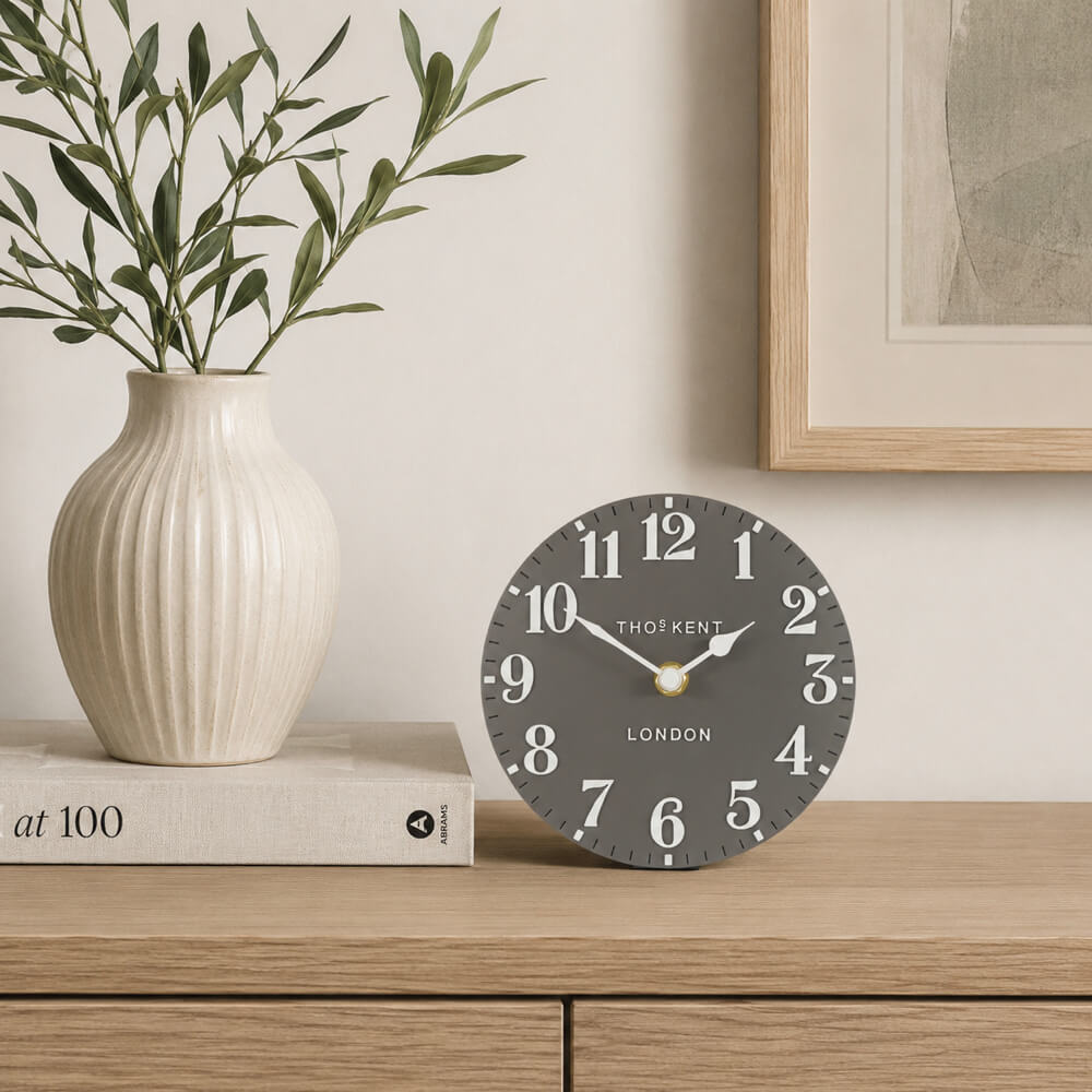 A round mantel clock with a dark grey face and white numerals, styled on a light wood console table next to a cream ribbed ceramic vase with green leaves.