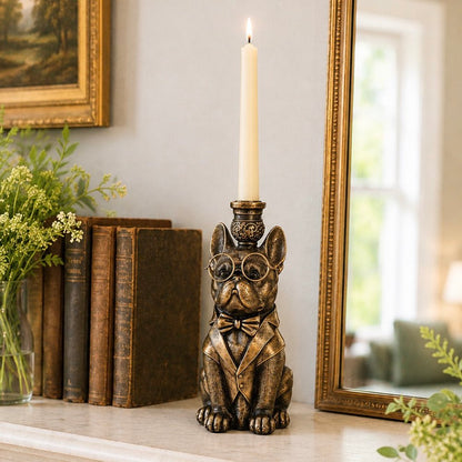 A bronze-finished French bulldog candle holder with round glasses, a bow tie, and a suit jacket, holding a lit white taper candle on a tabletop beside books and a mirror.