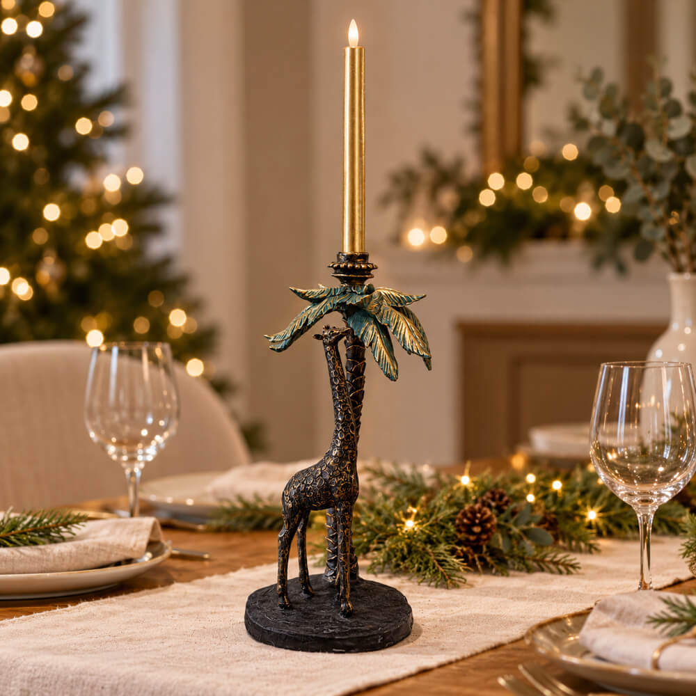 A decorative table centerpiece featuring a black giraffe holding a palm tree with a LED gold taper candle, set on a dining table with holiday greenery, plates, and wine glasses in a softly blurred festive interior background.
