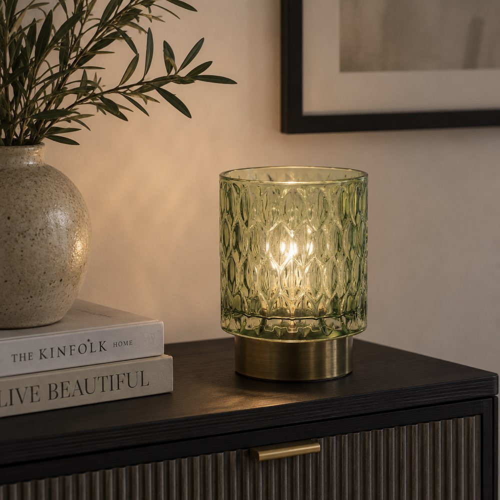 A green textured glass LED light with a warm glowing light inside, set on a dark wooden sideboard next to a vase and books in a styled interior setting.