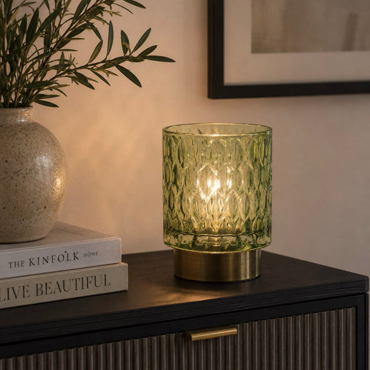 A green textured glass LED light with a warm glowing light inside, set on a dark wooden sideboard next to a vase and books in a styled interior setting.