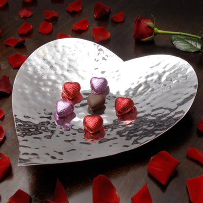 Heart-shaped silver dish with chocolate hearts surrounded by red rose petals on a wooden surface