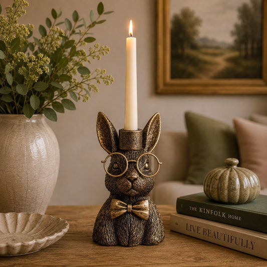 A decorative hare-shaped candle holder with a lit white candle, styled on a wooden table in a cosy living room setting with a vase, framed art, and books.