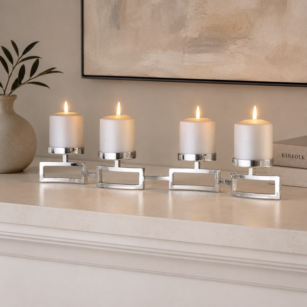 Four white pillar candles in a row on a silver metal multi-candle holder with a polished rectangular base, set on a light cream mantelpiece or shelf with a vase and framed art in the background.