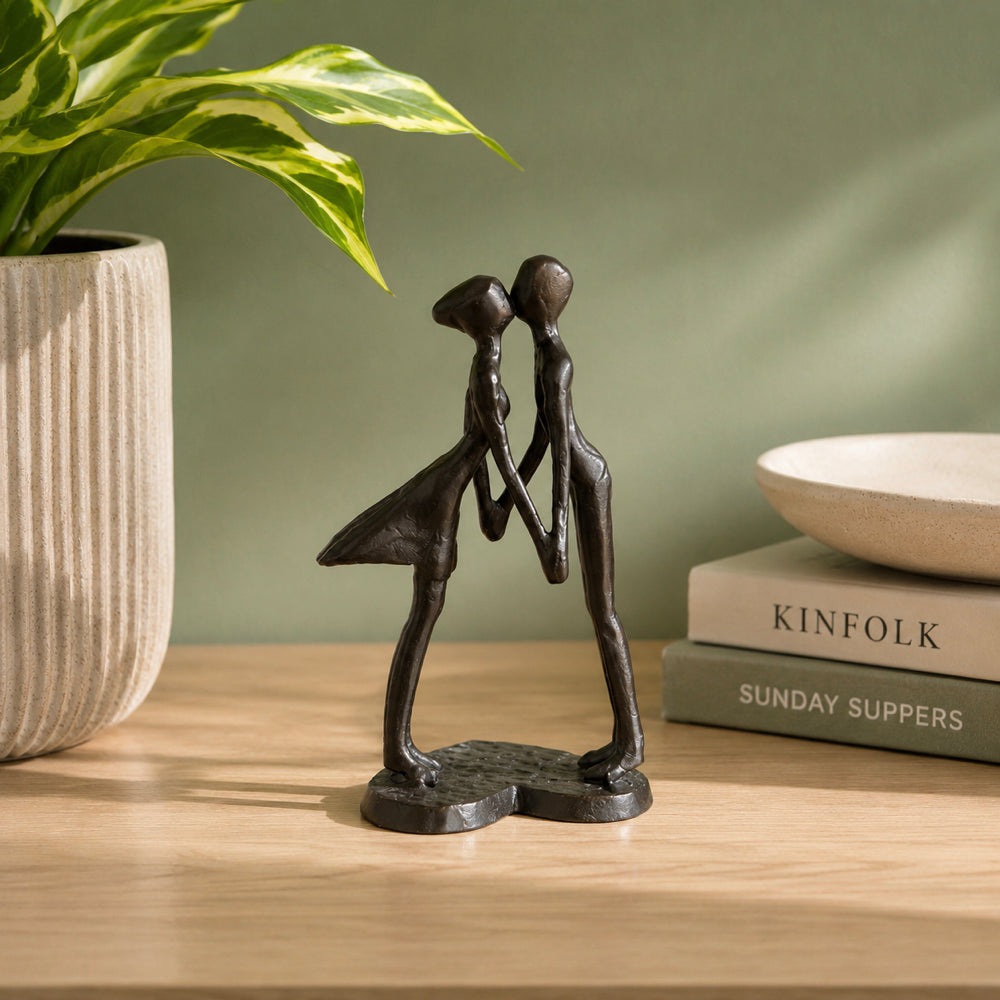 A small dark bronze figurine of two abstract figures kissing stands on a light wood tabletop. A potted plant is on the left, and stacked books with a shallow bowl are on the right against a muted green wall.