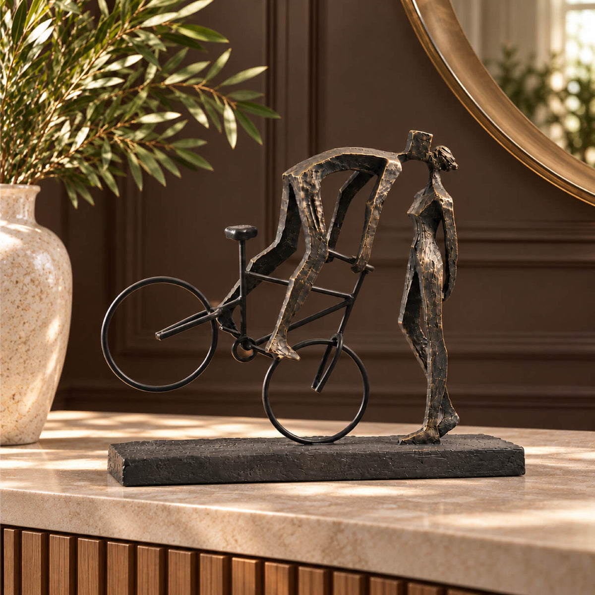 A dark metal sculpture of a cyclist kissing a lady against a standing figure, displayed on a light stone console table in a warmly lit interior setting.