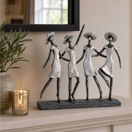 Four dancing women figurine sculpture in a row, featuring textured silver and black finishes, displayed on a mantelpiece in a home setting next to a lit candle and a potted plant.
