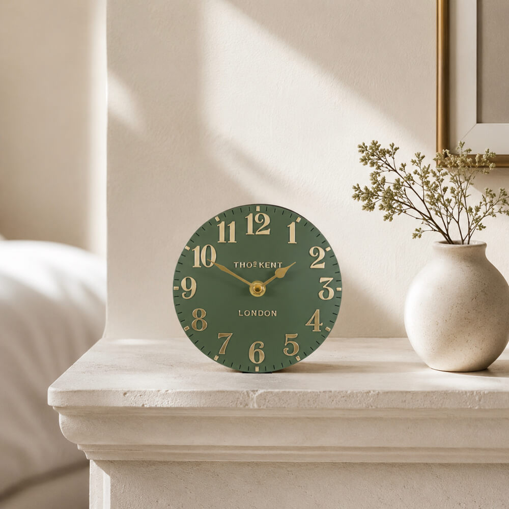 A round green mantel clock with gold numerals and hands sits on a stone mantelpiece in a softly lit room, next to a small beige vase with dried stems.