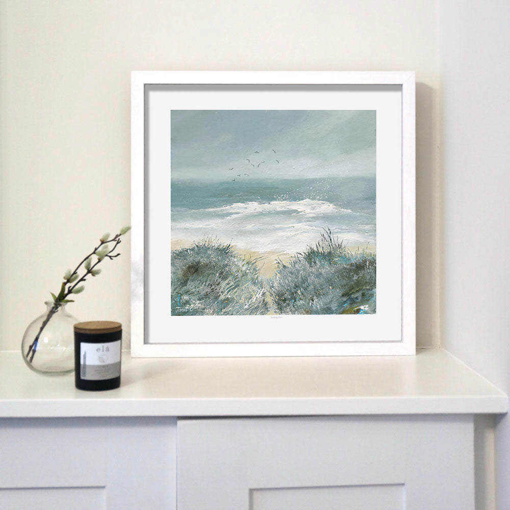 A framed print titled 'Looking Out' depicting a serene ocean scene with soft, muted tones and expressive brushstrokes, placed on a white surface, with a candle to the left side of the frame.
