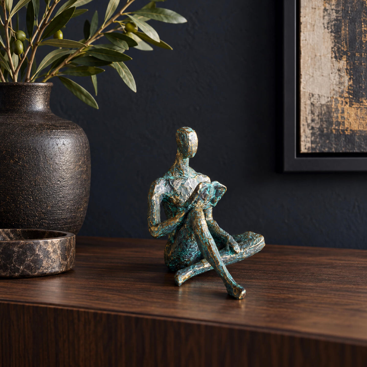 A small abstract figurative sculpture of a seated person with a textured turquoise and bronze finish, displayed on a dark wood mantel next to a dark ceramic vase with greenery.