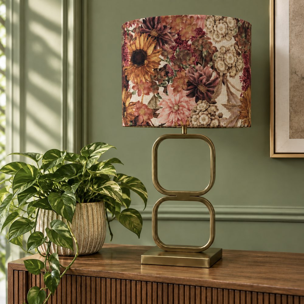A table lamp with a cylindrical drum shade featuring a vintage floral pattern in warm tones, set on a wooden console table next to a potted green plant against a sage green wall.