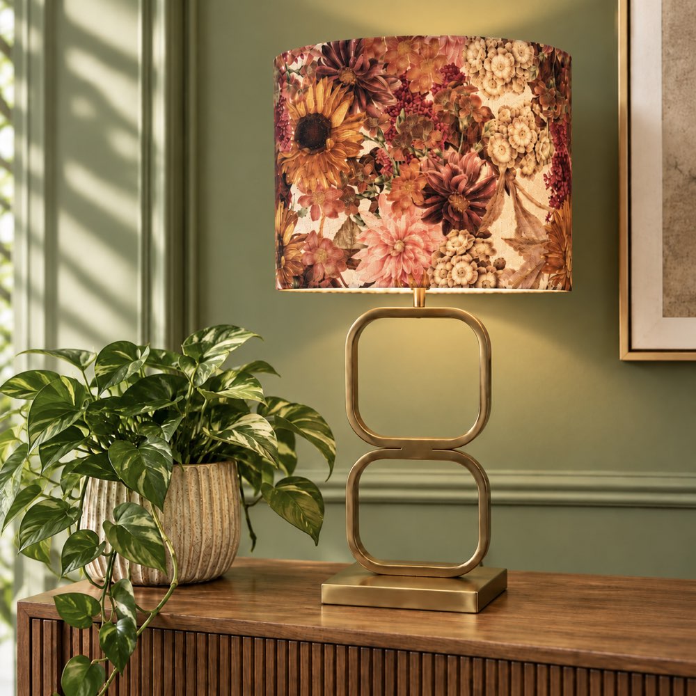 A table lamp with a cylindrical floral drum shade in warm autumn tones and a gold-finished base featuring two stacked square loops, styled on a wooden console table next to a green potted plant against a sage green wall.