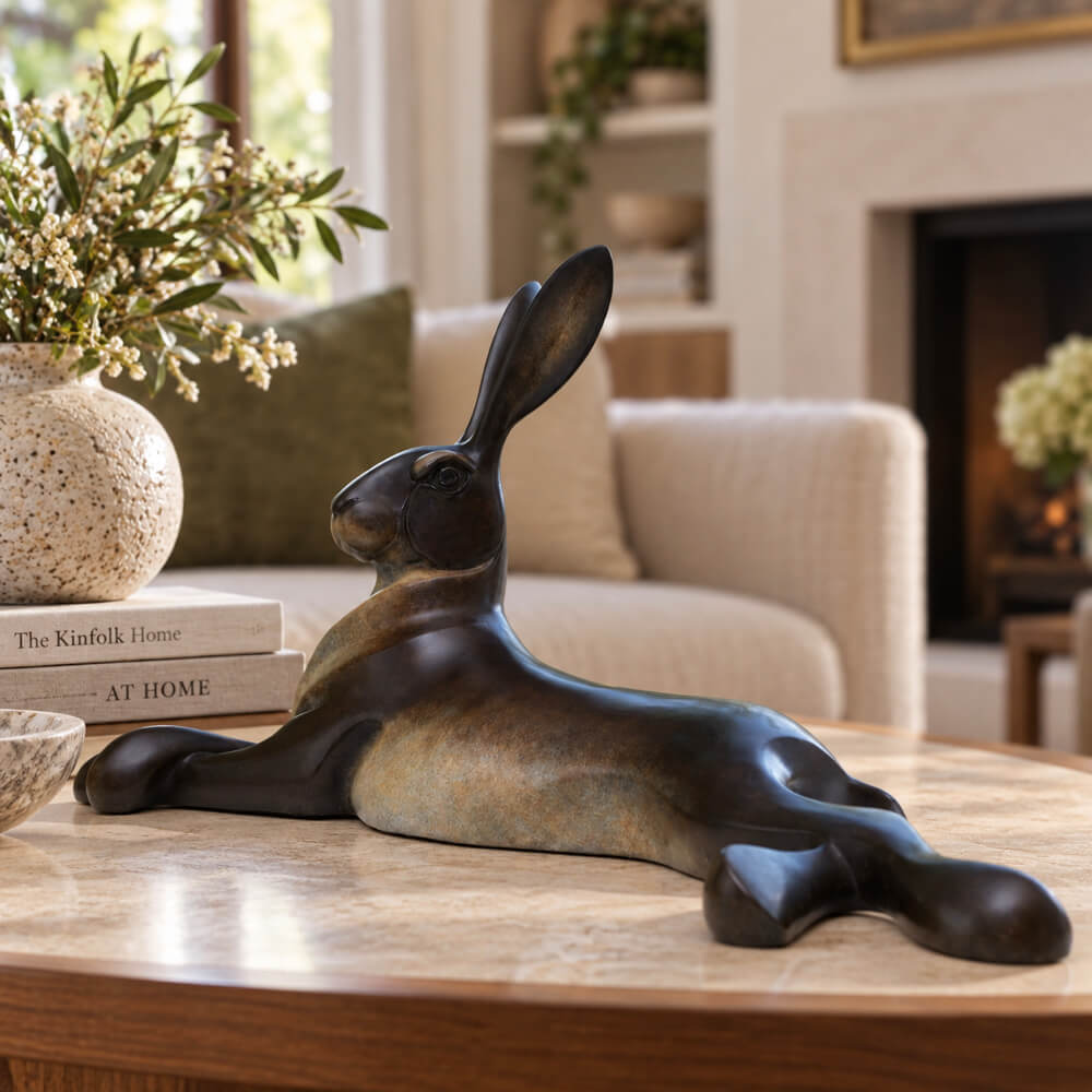 A dark bronze-finished sculpture of a reclining rabbit resting on a round wooden coffee table in a cozy living room setting.