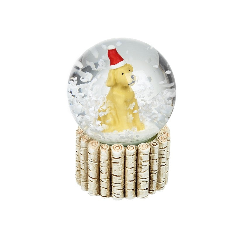 A decorative snow globe figurine featuring a small yellow dog wearing a red Santa hat, surrounded by white faux snow inside a clear globe, set on a base made of light wood logs, isolated on a white background.
