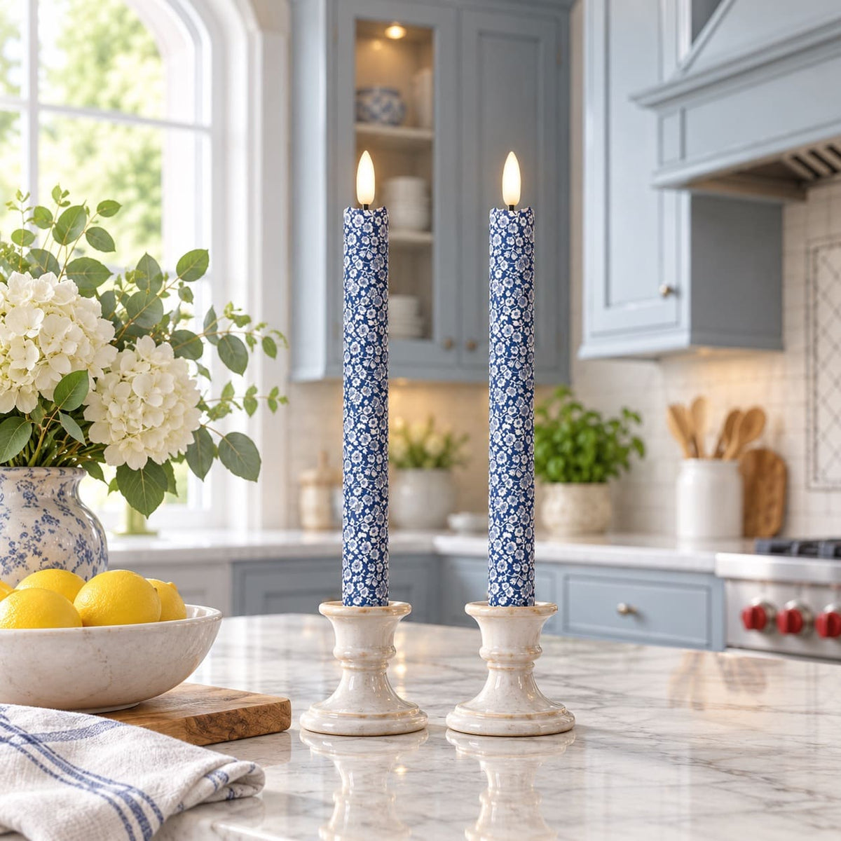 Two tall blue-and-white patterned LED taper candles in white candleholders on a marble kitchen counter, with flowers, lemons, and blue cabinets in the background.