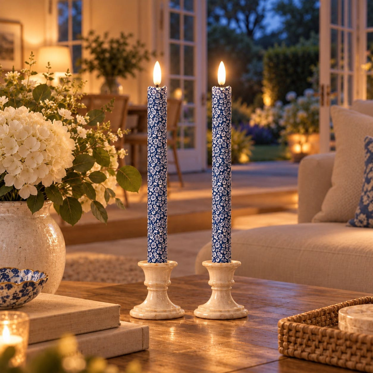 Two tall blue-and-white patterned LED taper candles in stone candleholders, lit and displayed on a wooden table in a warmly lit living room with flowers and a garden view in the background.