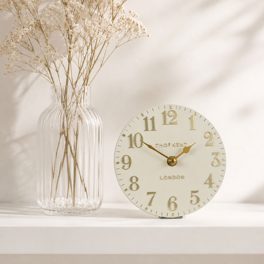 A round mantel clock with a cream-coloured face and gold hands, styled next to a ribbed clear glass vase with dried white flowers on a white shelf against a softly shadowed background.