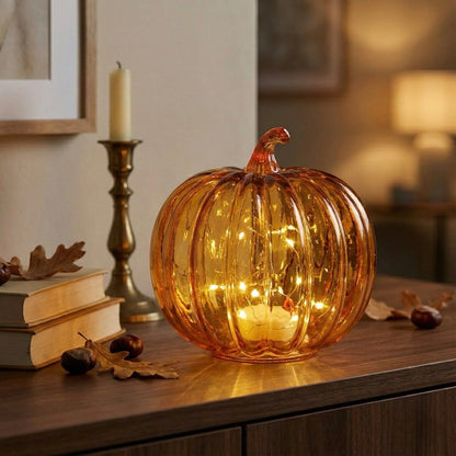 An amber glass pumpkin-shaped decorative light with warm white fairy lights inside, placed on a wooden table in a cozy indoor setting with books, a candle, and acorns nearby.