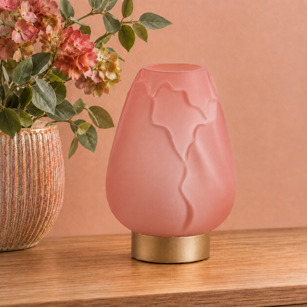 A pink sculptural table lamp with a brushed gold base, styled on a wooden surface next to a vase of flowers against a peach-colored background.