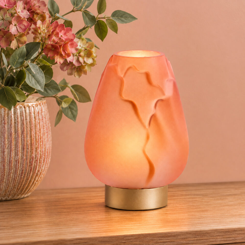 A glowing peach-orange table lamp with a sculpted embossed design on its shade and a gold-toned circular base, placed on a wooden surface next to a vase of pink flowers against a warm beige wall.