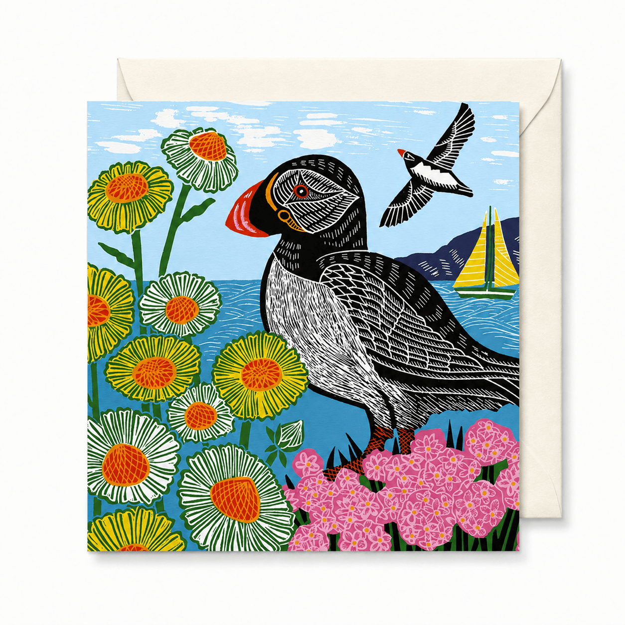 A square illustrated greeting card with a cream envelope behind it. The card artwork shows a black-and-white puffin with a red-orange beak standing among yellow, white, and pink flowers, with blue water and sky, a small sailboat, and another bird in flight.