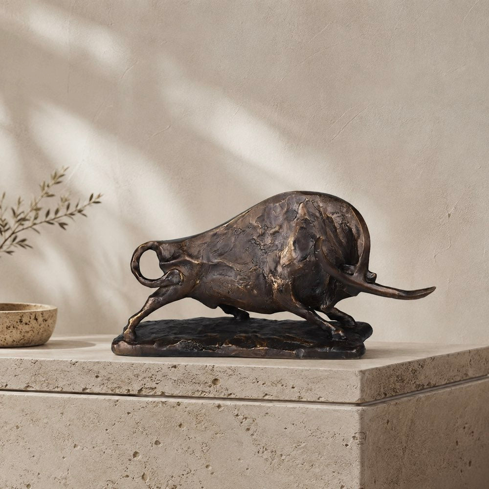 A bronze resin bull sculpture in a dynamic charging pose, displayed on a stone pedestal against a textured beige wall with soft leaf shadows.