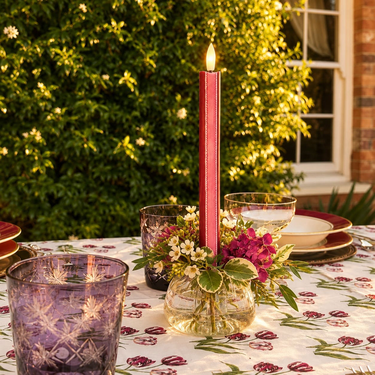 A red and pink striped LED taper candle stands in a clear glass vase on a decorated outdoor table, surrounded by purple glassware, plates, and a floral tablecloth, with a green garden and window in the background.