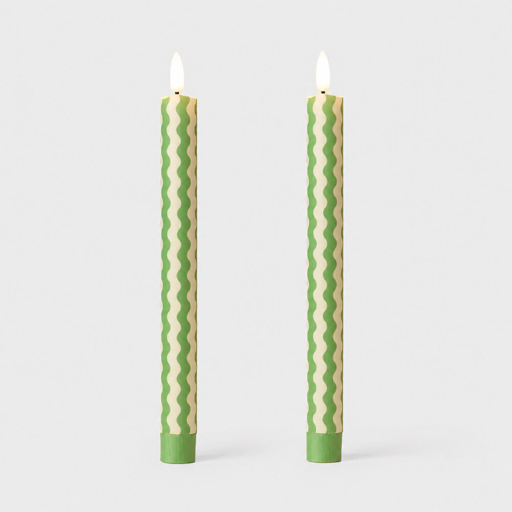 Two tall taper candles with a wavy striped pattern in light green and cream, shown standing upright on a light grey background with their flames lit.