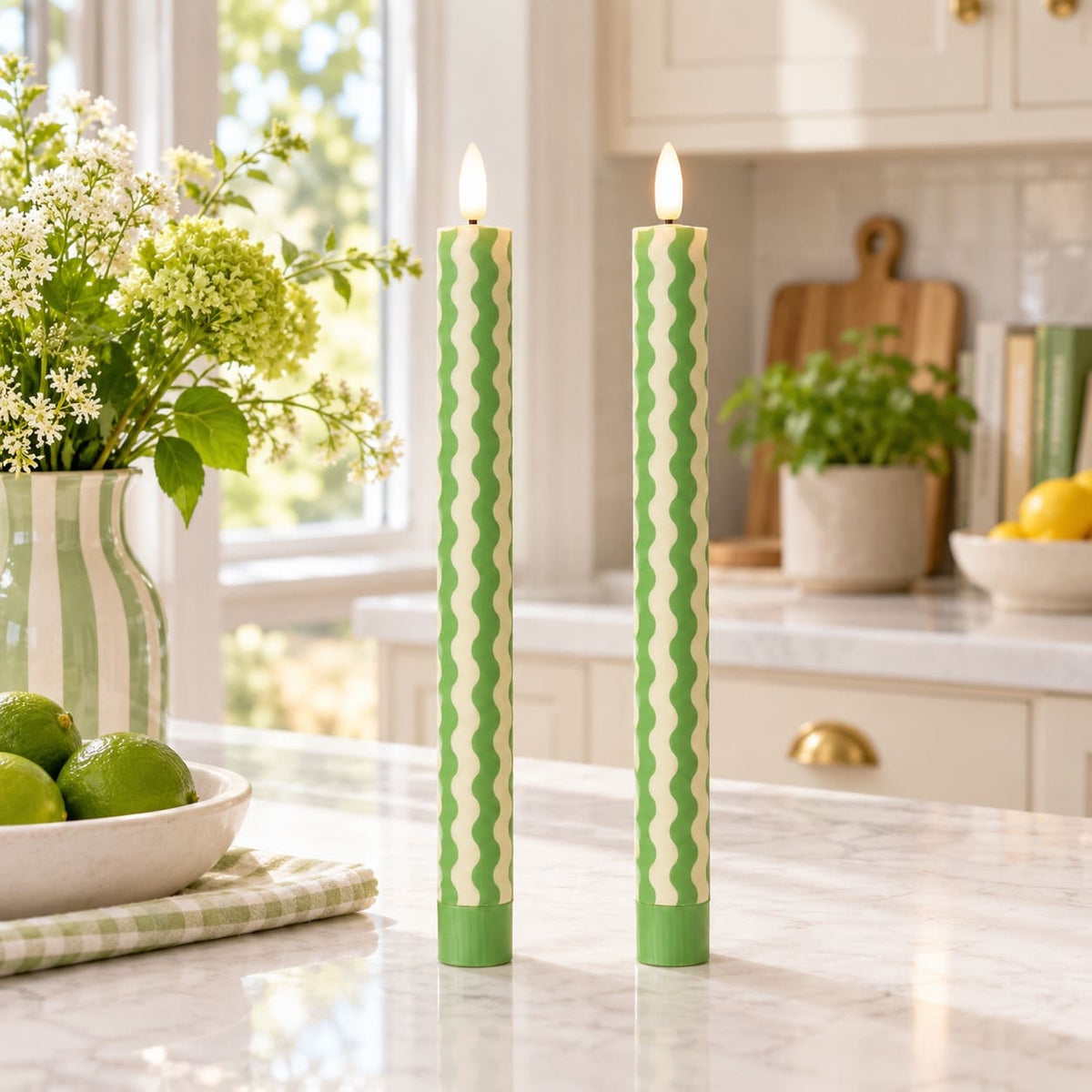 Two tall green and cream wavy patterned candles standing on a marble kitchen counter, with flowers, limes, and a blurred kitchen background.