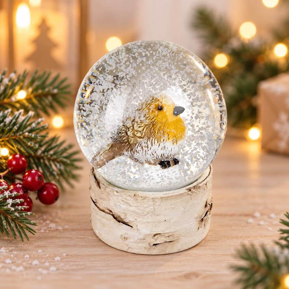 A Christmas-themed snow globe featuring a small robin bird in a snowy scene, set on a birch wood base with festive pine branches and warm string lights in the background.