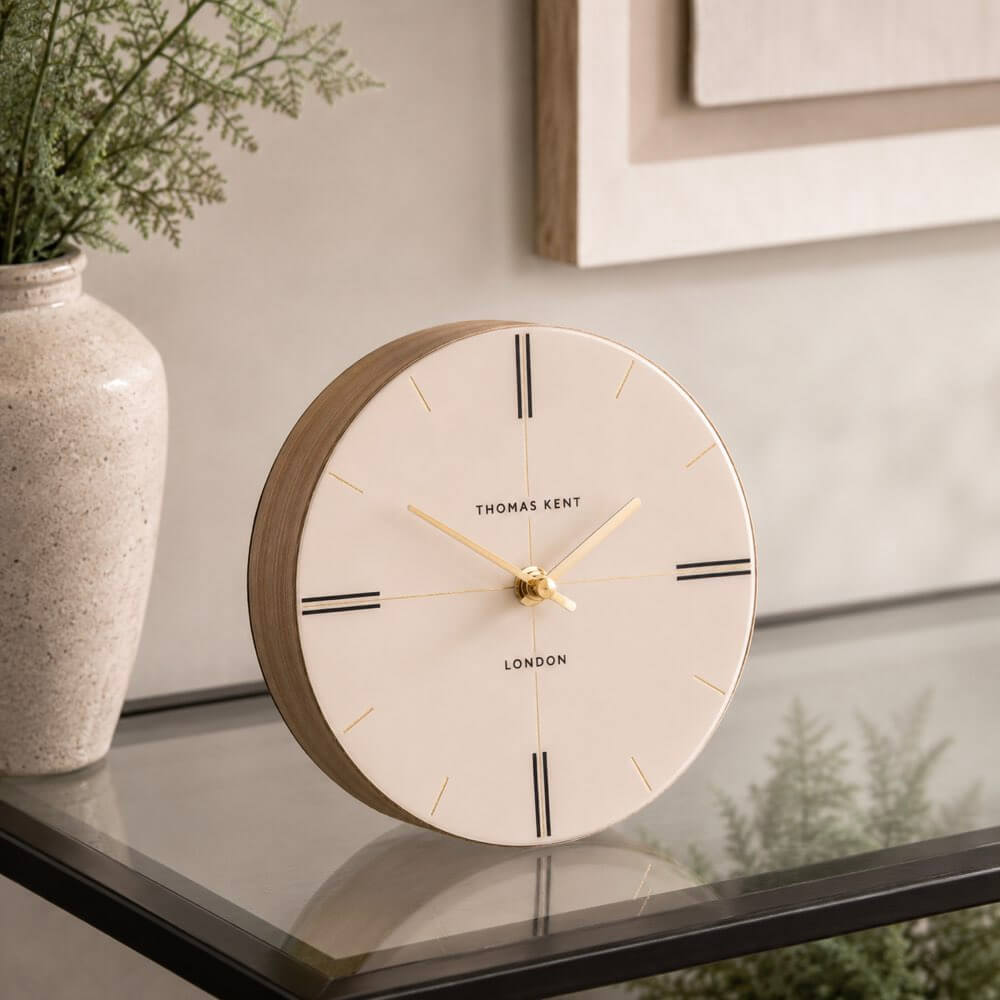 Round clock with wooden frame on a surface, next to a vase with plants.