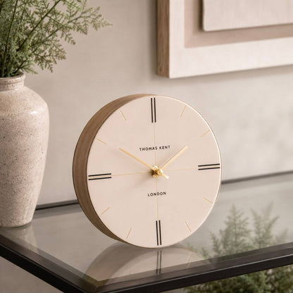 Round clock with wooden frame on a surface, next to a vase with plants.