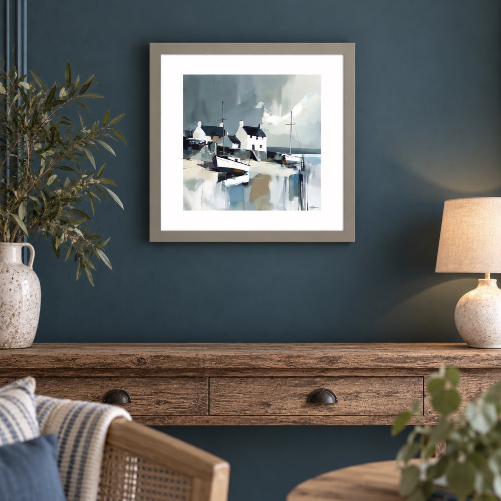 A framed art print depicting a coastal village with small boats and houses, displayed on a dark teal wall above a rustic wooden console table with a lamp and vase.