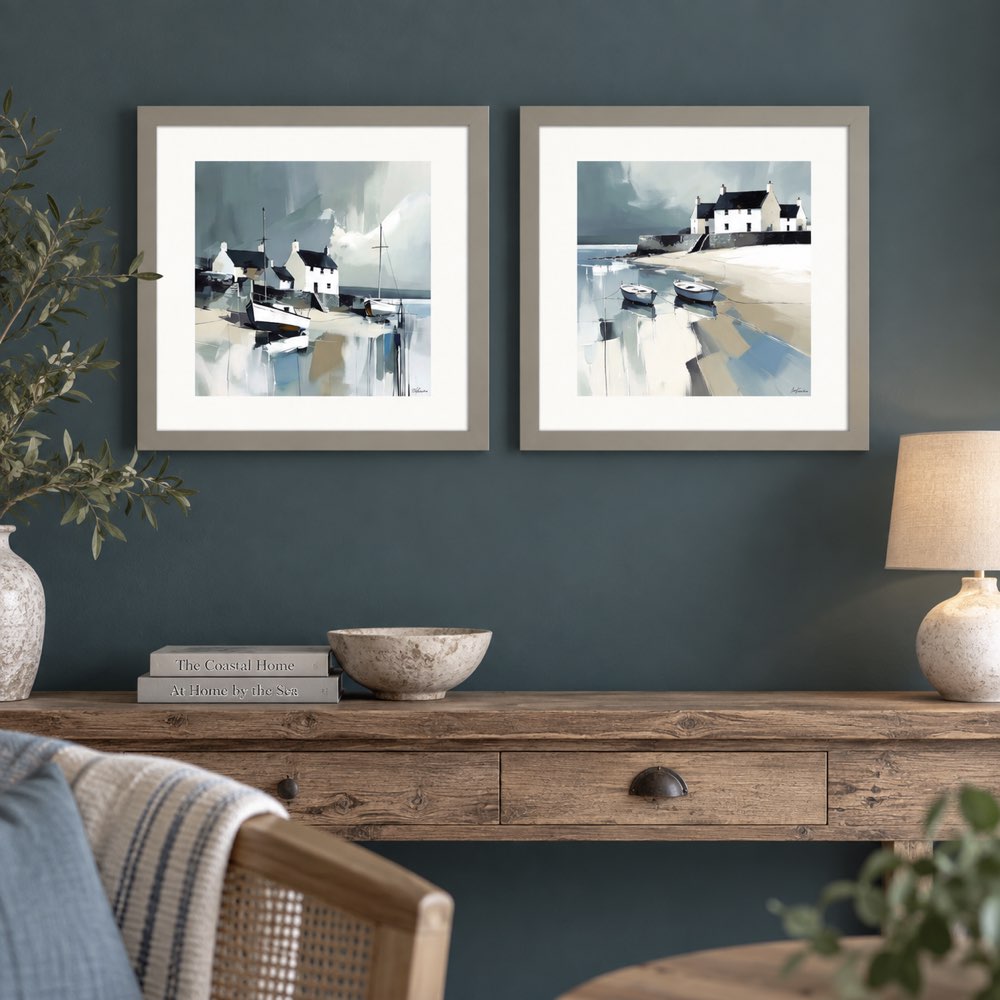 Two framed coastal landscape prints hang side by side on a dark teal wall above a rustic wooden console table. The prints depict small boats, white houses, and icy water in soft blue, gray, and white tones.
