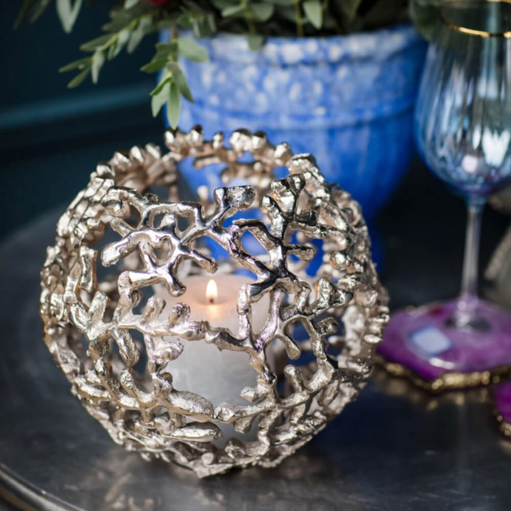 A spherical silver coral-like metal candle holder with a lit tealight candle inside, styled on a dark tabletop with a blue vase and glassware in the background.