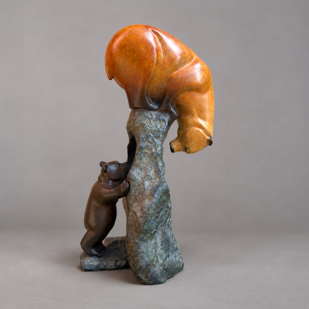 A bronze sculpture of two bears, one large bear climbing a tall bronze rock while a smaller bear stands below, photographed against a neutral grey studio background.