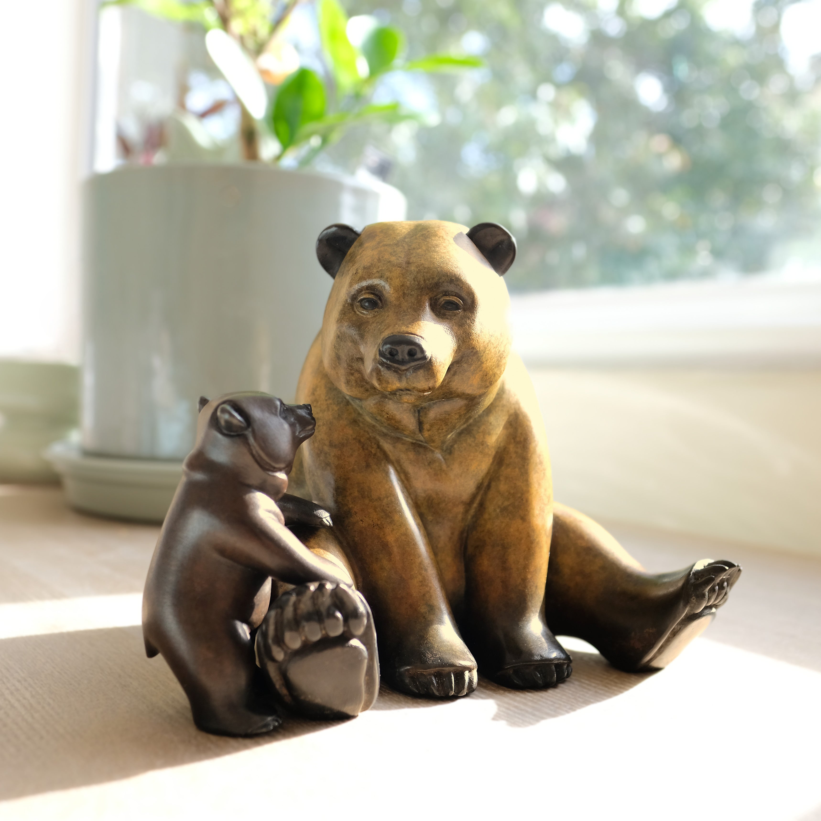 Bronze statue of a bear and a cub on a light surface with a blurred plant and window in the background.