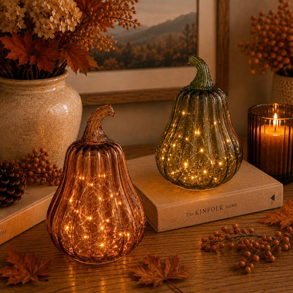 Two ribbed glass decorative pumpkins with warm white fairy lights inside, styled on a wooden table with autumn leaves, a candle, books, and dried floral arrangements in a cozy indoor setting.