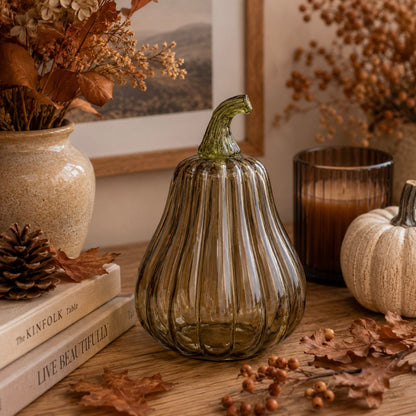 A ribbed glass pumpkin vase in smoky olive green sits on a wooden table surrounded by autumn decor, including dried leaves, a small white pumpkin, books, pinecone, and a candle.
