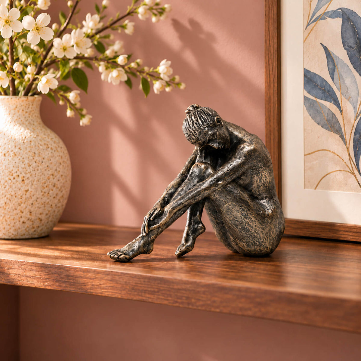The Contemplating Woman Sculpture on a walnut wooden shelf against a dusty pink wall, styled with white blossom flowers.